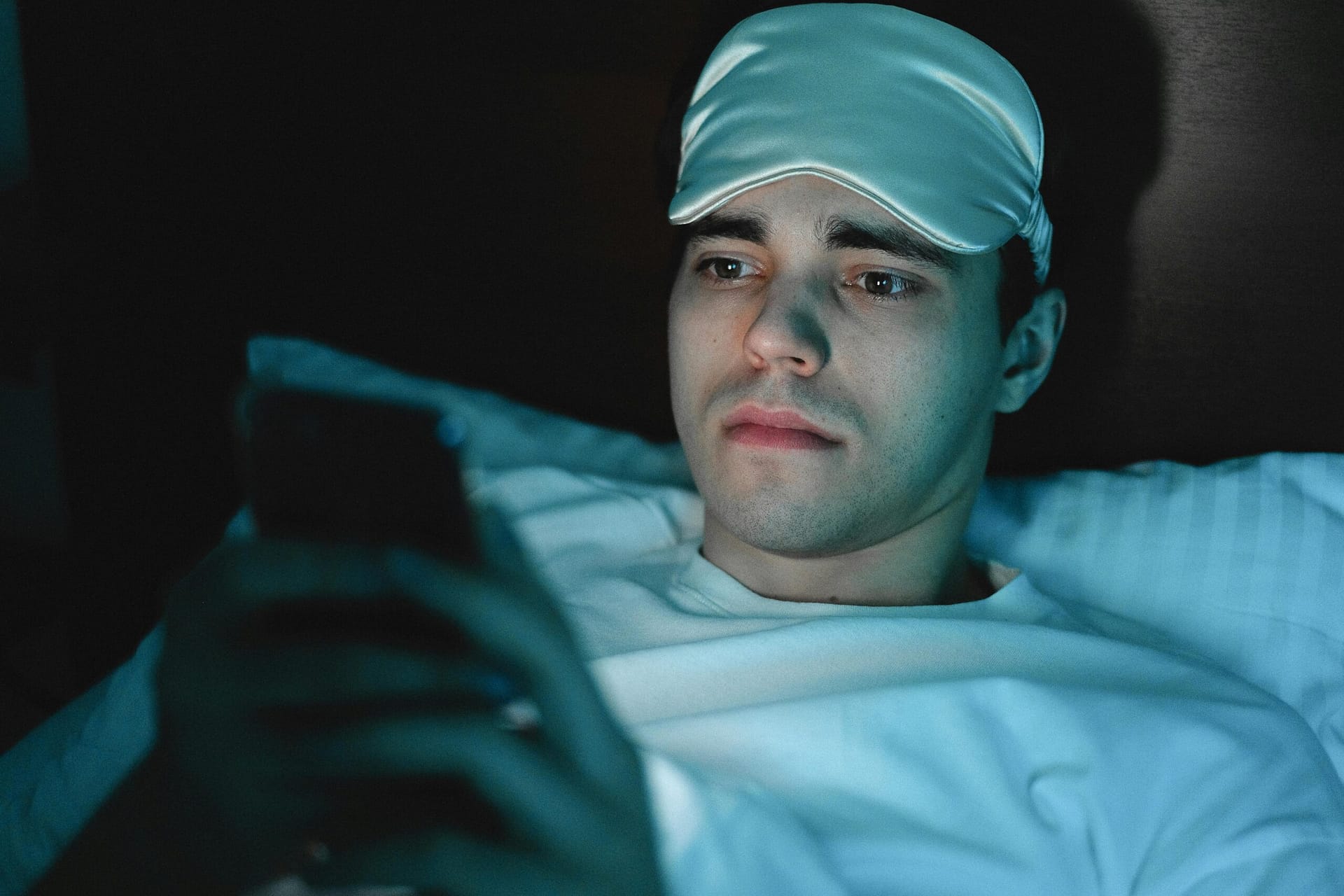 A person using a smartphone in bed at night, blue light glowing on their face, illustrating sleep disruption.