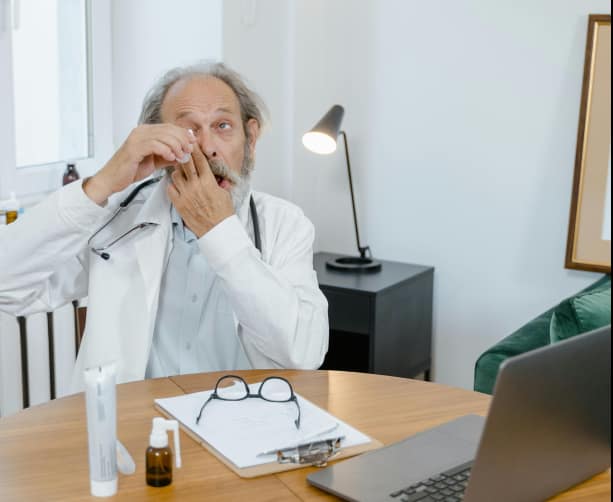 Person using eye drops at a desk with a water bottle, symbolizing relief from dryness through hydration.